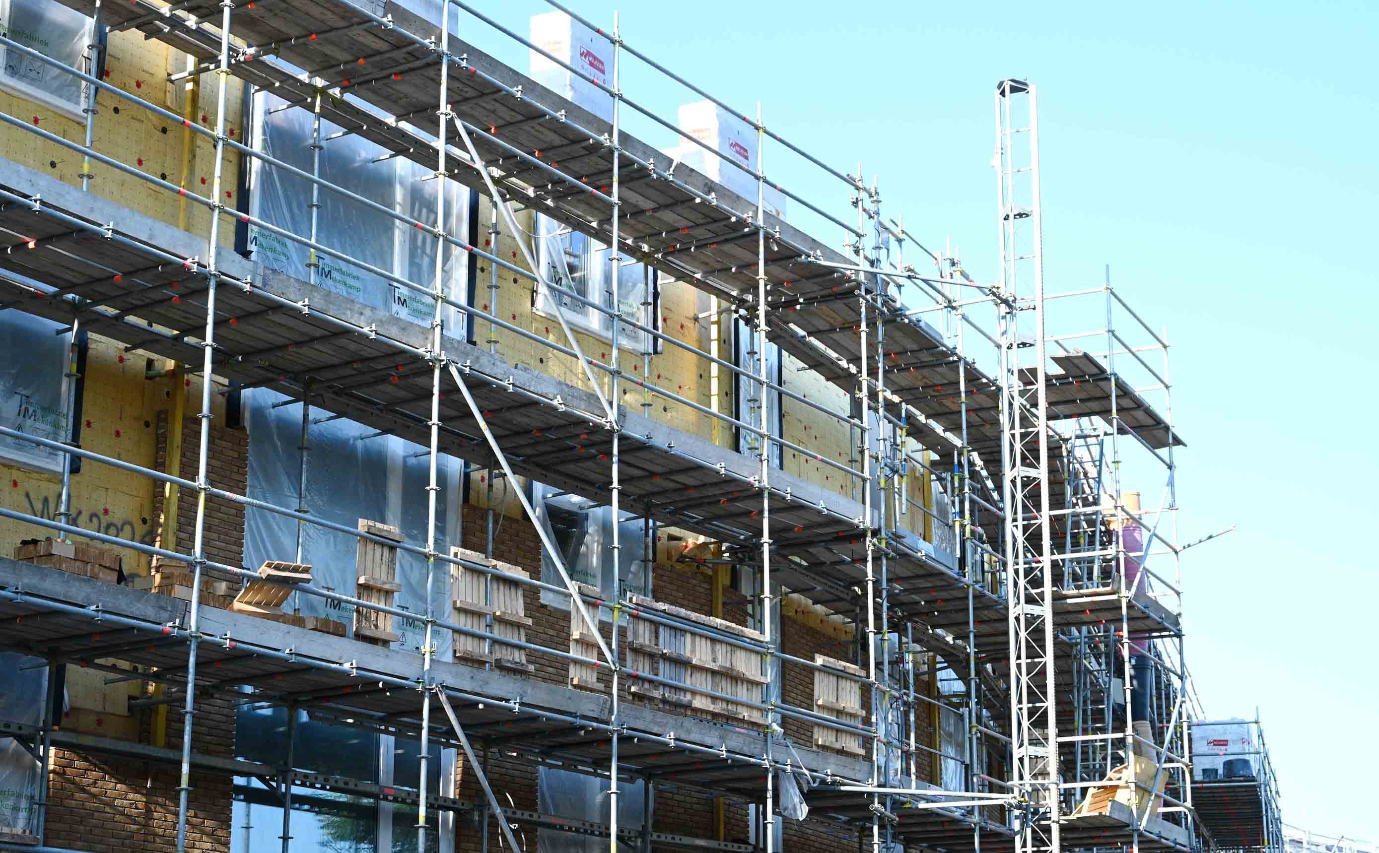 Yellow mineral wool insulation batts being fitted to a facade under construction, wrapped in white membrane, with a brick tower and scaffolding behind it against a clear sky.
