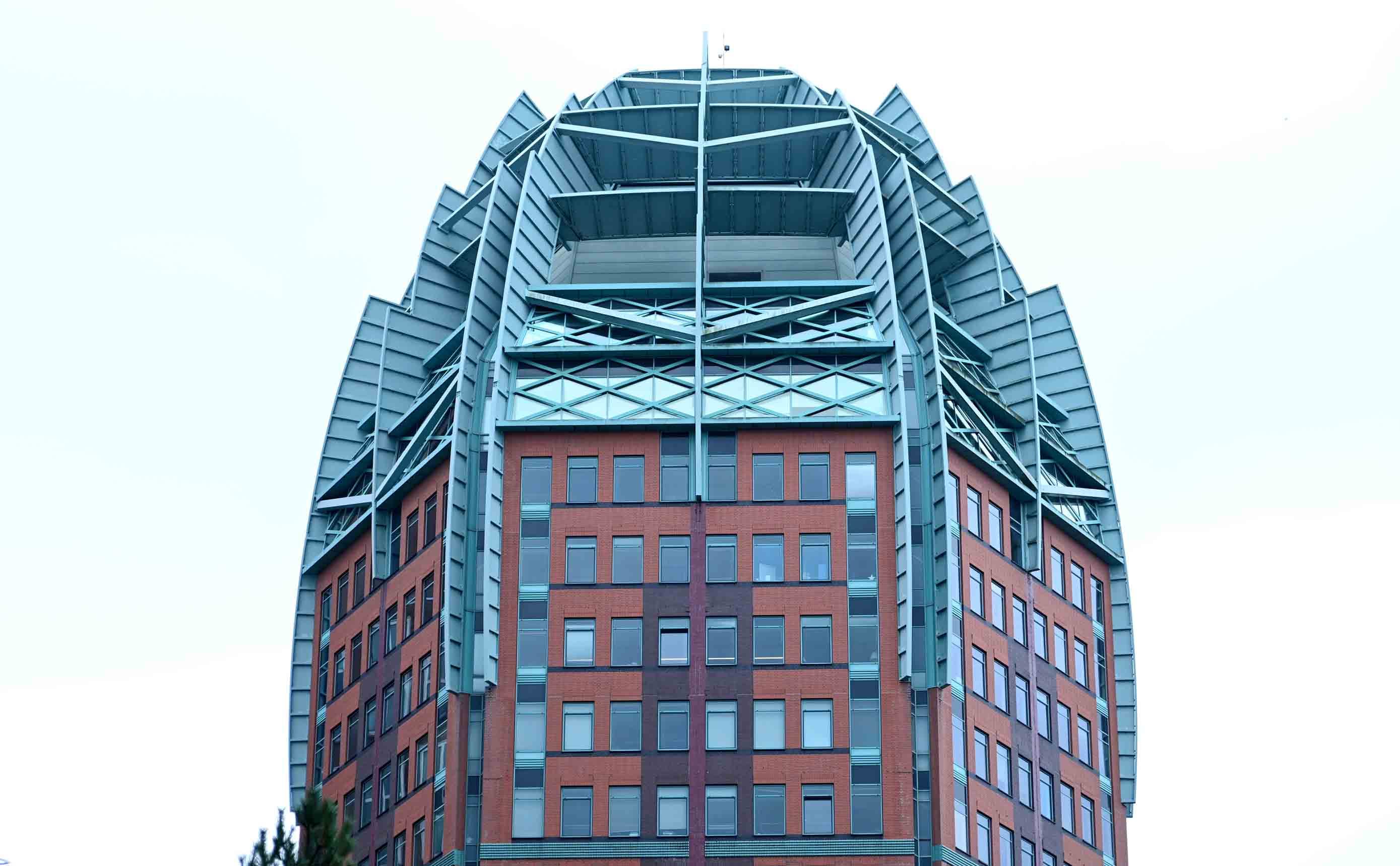A contemporary brick office tower topped by a curved steel and glass crown, its structural frame, brick skin and glazing all visible at once against a pale sky.