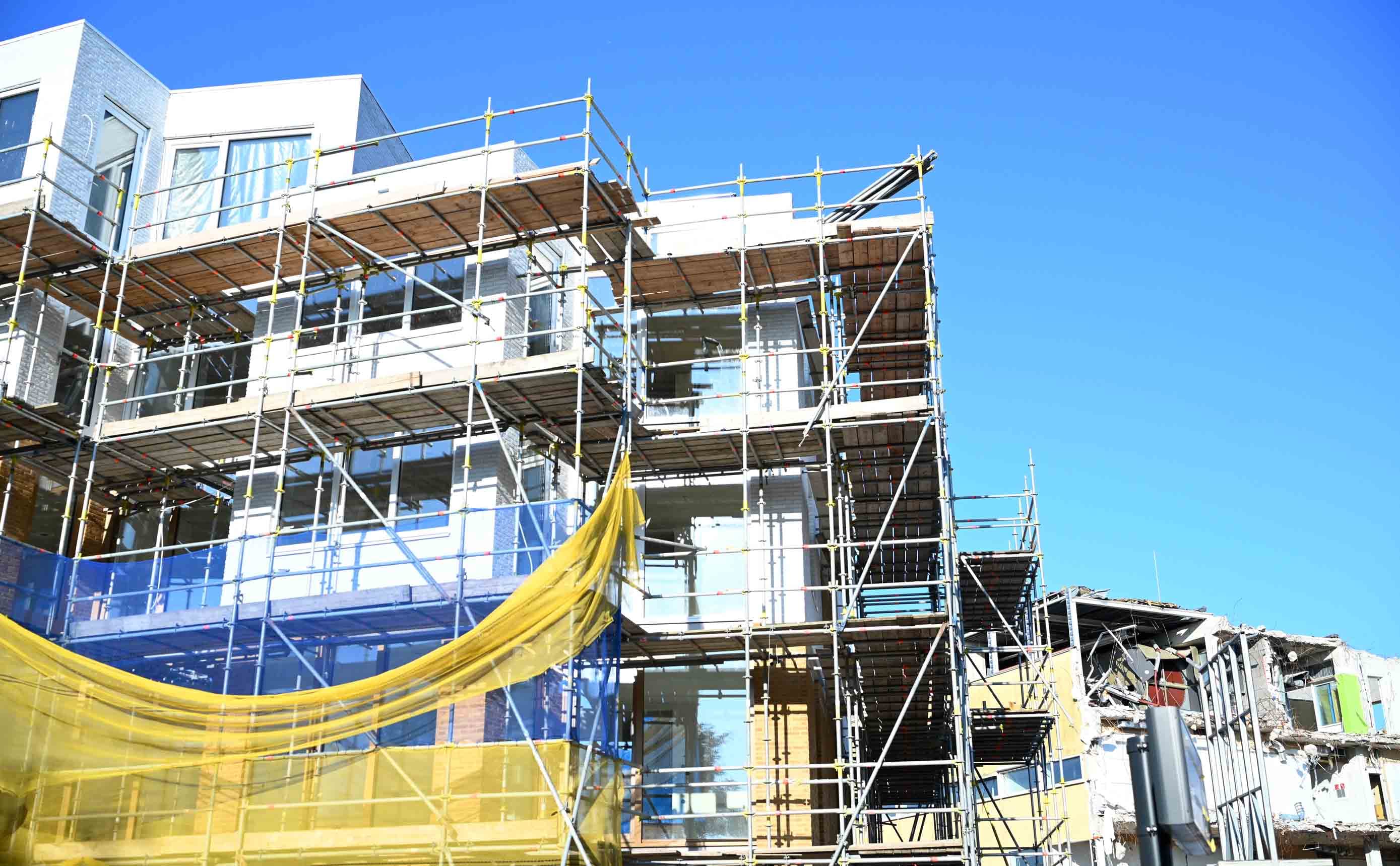 A residential building under construction wrapped in scaffolding, with a brick outer leaf, yellow mineral-wool insulation, glazing and precast panels being fitted to the facade against a clear blue sky.