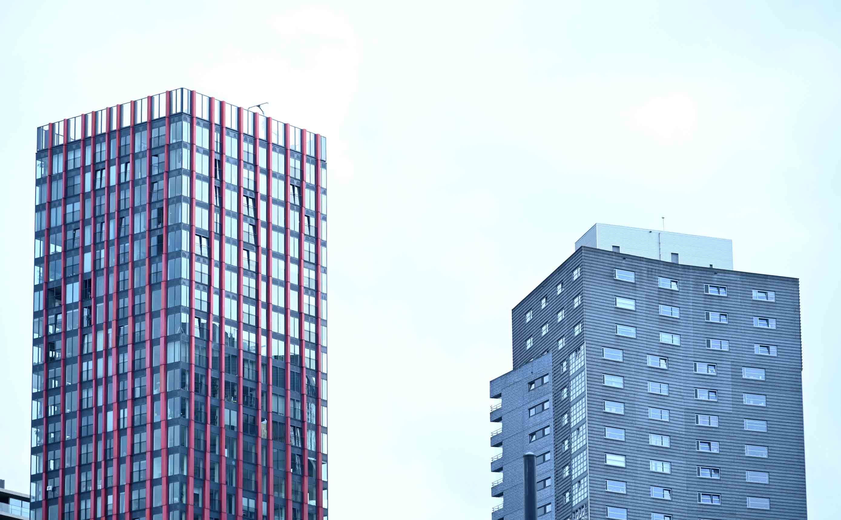 Twee opgeleverde woontorens in Nederland tegen een lichte lucht, een met rood en glas bekleed en een in grijze baksteen, allebei af en in gebruik.
