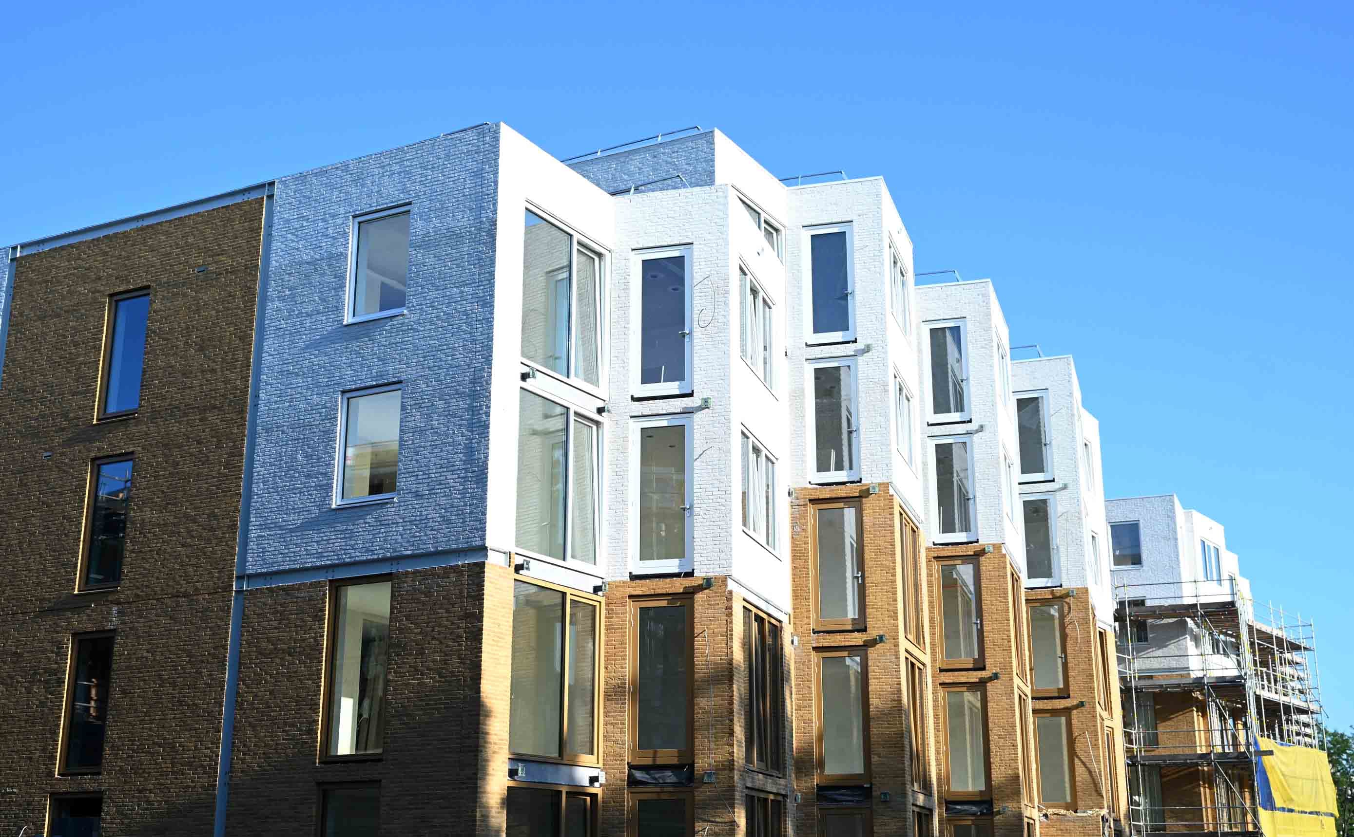Een rij net opgeleverde Nederlandse appartementen onder een strakblauwe lucht, met bakstenen gevels in grijs, wit en warm bruin, grote glazen erkers, en aan het einde nog steigers.