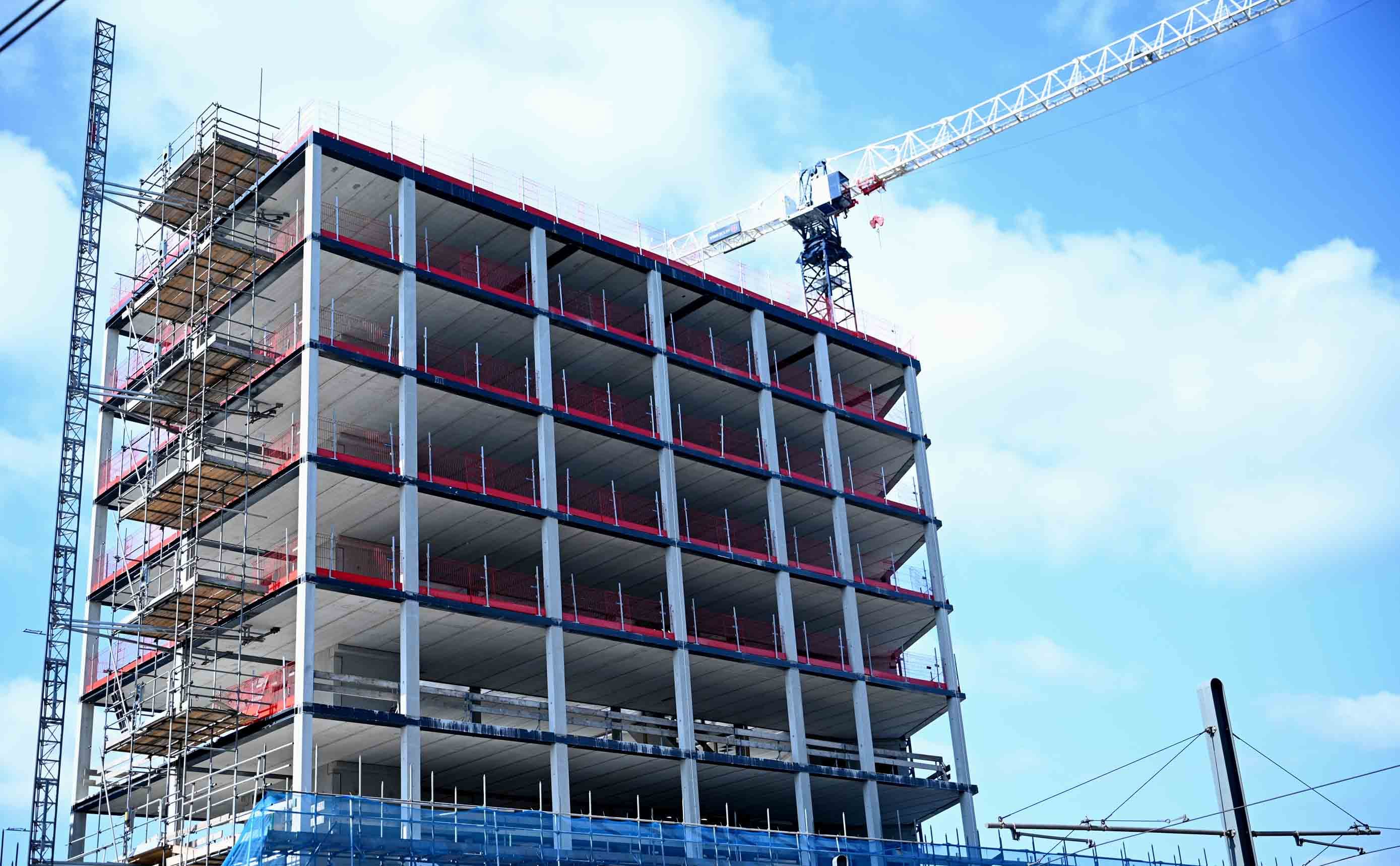 A multi-storey concrete building frame under construction, floor slabs and columns exposed, with a tower crane against a blue sky.