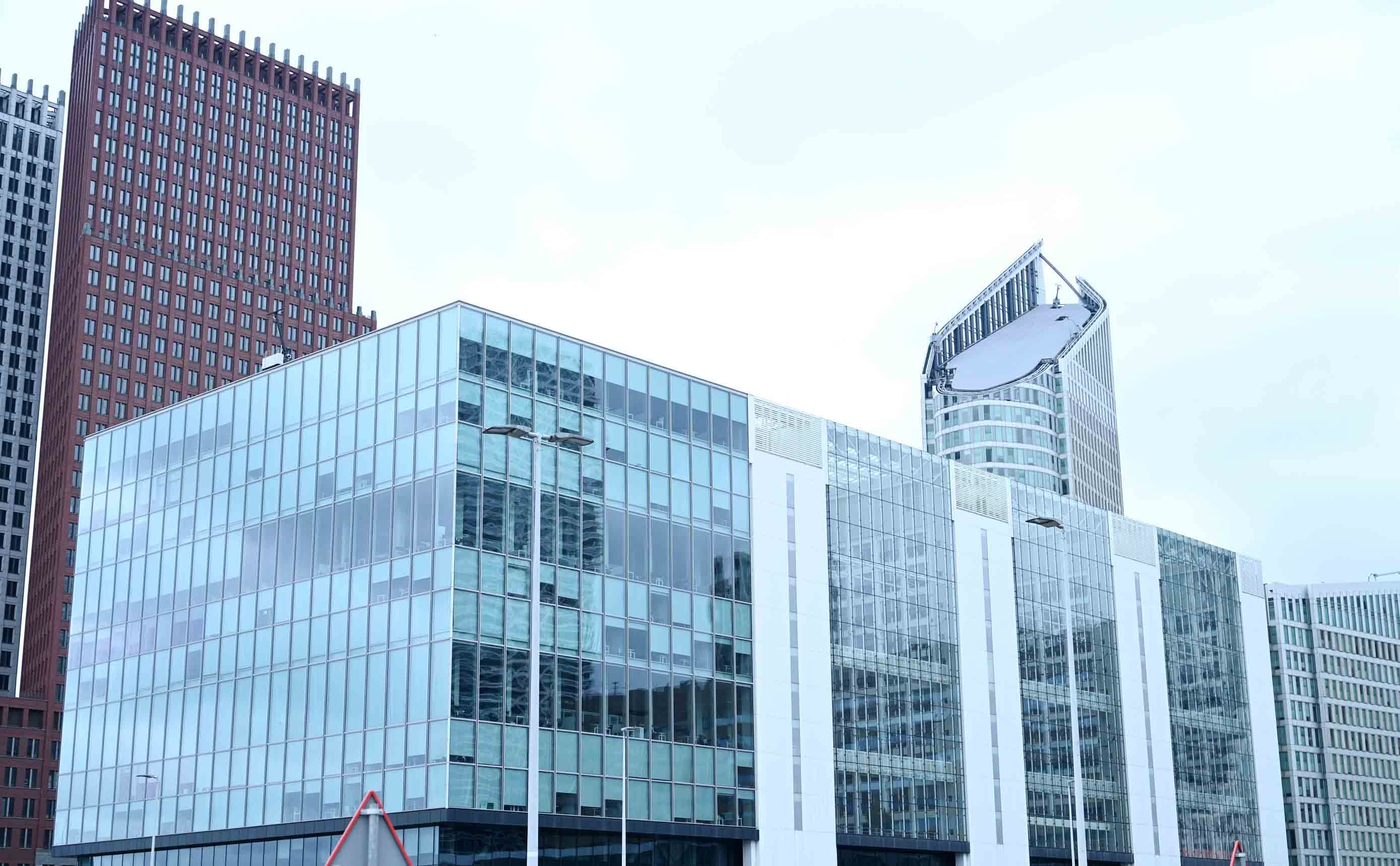 A large modern office building clad in a regular grid of glass curtain-wall panels, with taller brick and glass towers behind it against a pale sky, every floor and bay clearly repeated.