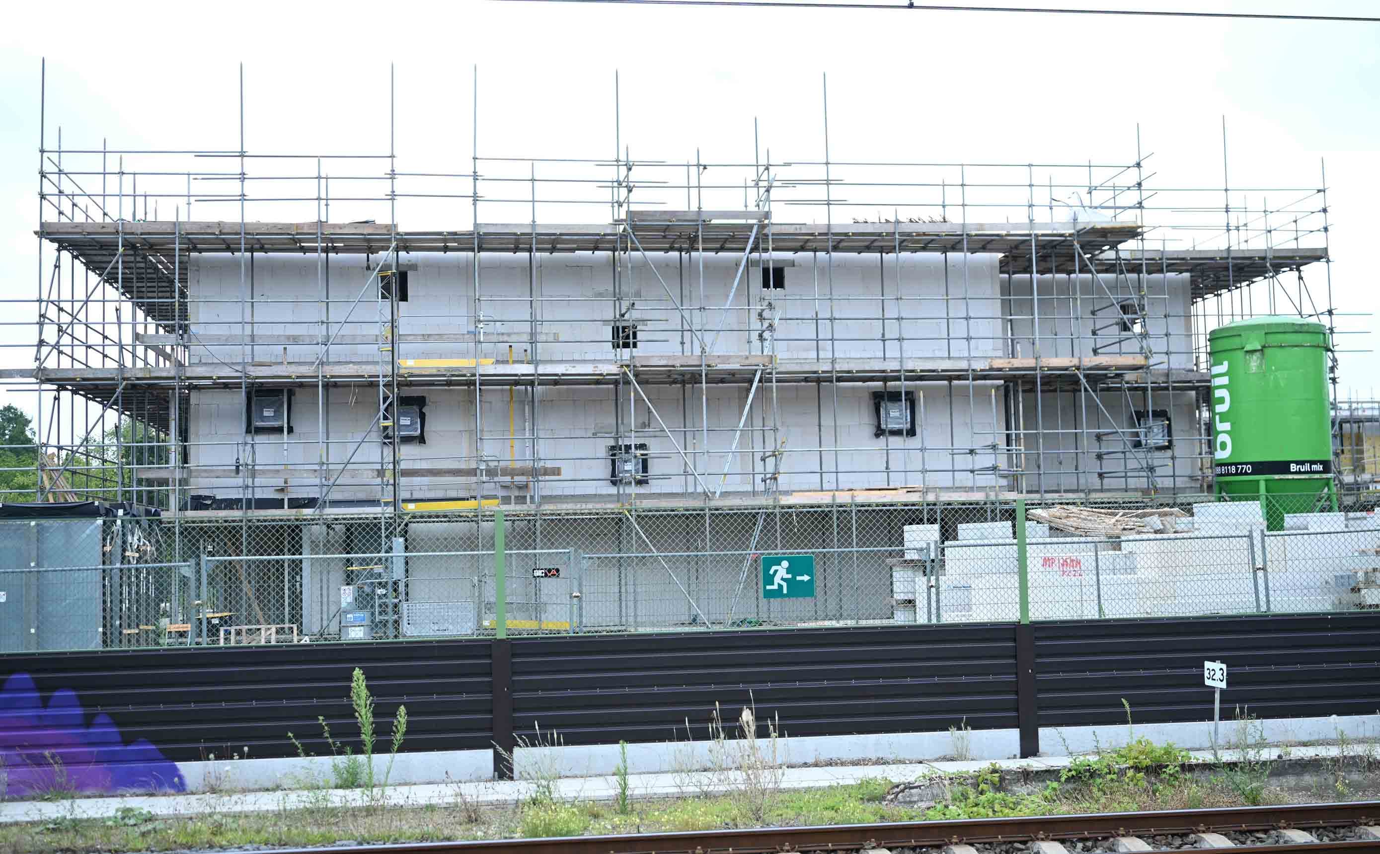 Een woongebouw in aanbouw met een betonnen skelet tegen een blauwe lucht, de verdiepingen nog open met rode randbeveiliging en een torenkraan naast de onafgebouwde ruwbouw.