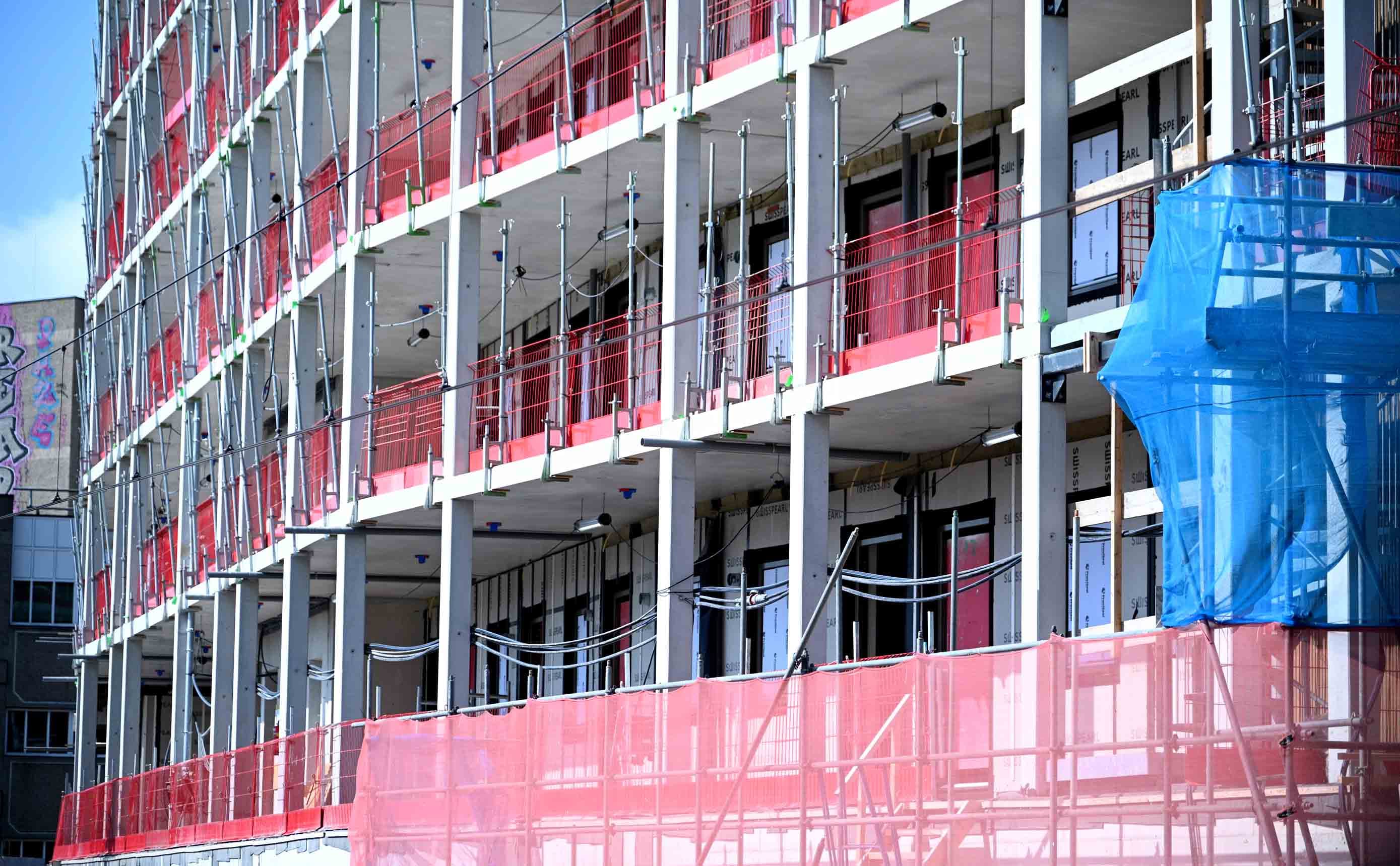 Een woongebouw in aanbouw, met de betonnen constructie en vloeren open in de lucht, raamopeningen en rode randbeveiliging langs elke verdieping.