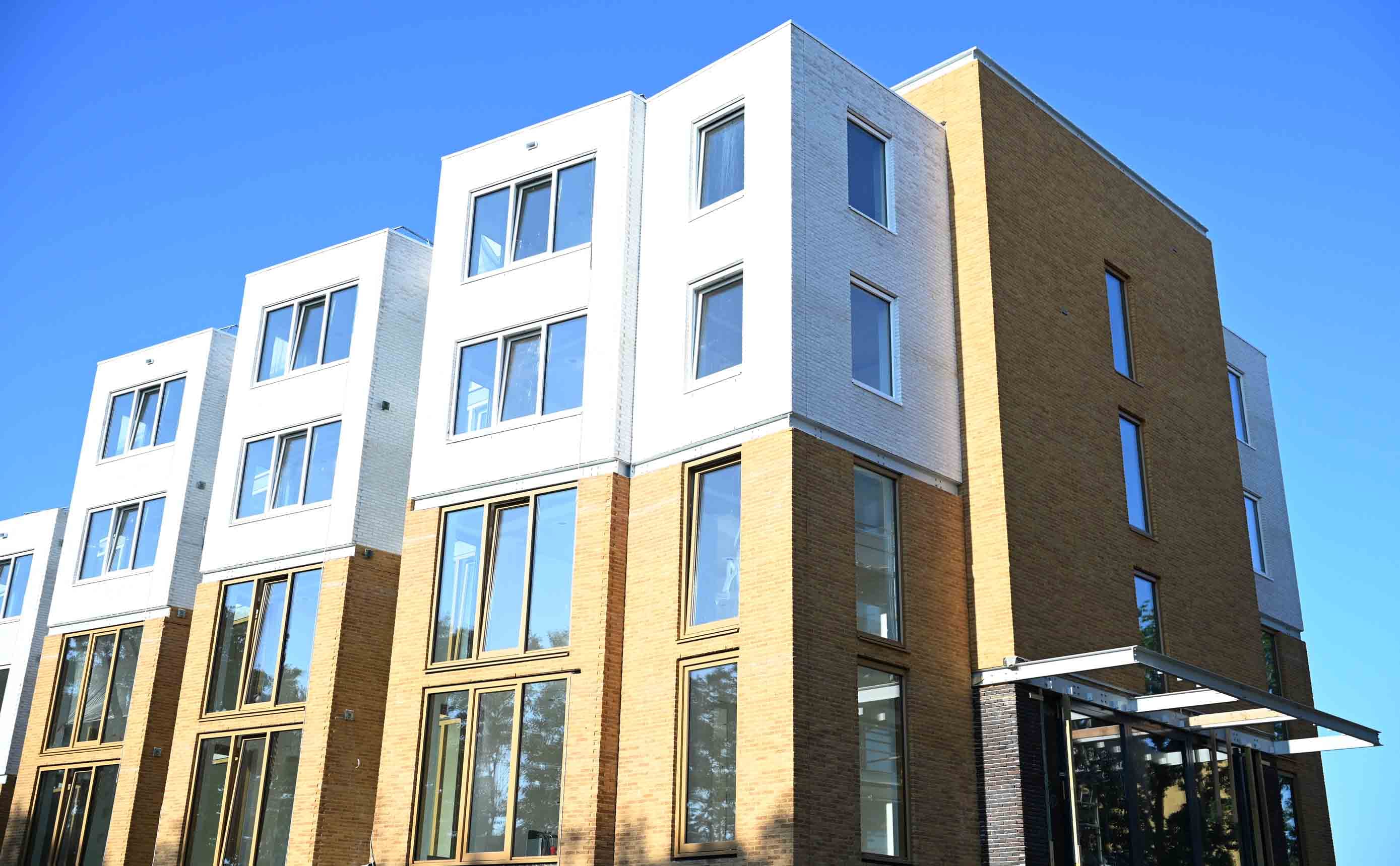 A completed contemporary Dutch apartment building of four storeys, facades in yellow-brown brick and white render, with large windows and a glazed ground-floor entrance, under a clear blue sky.