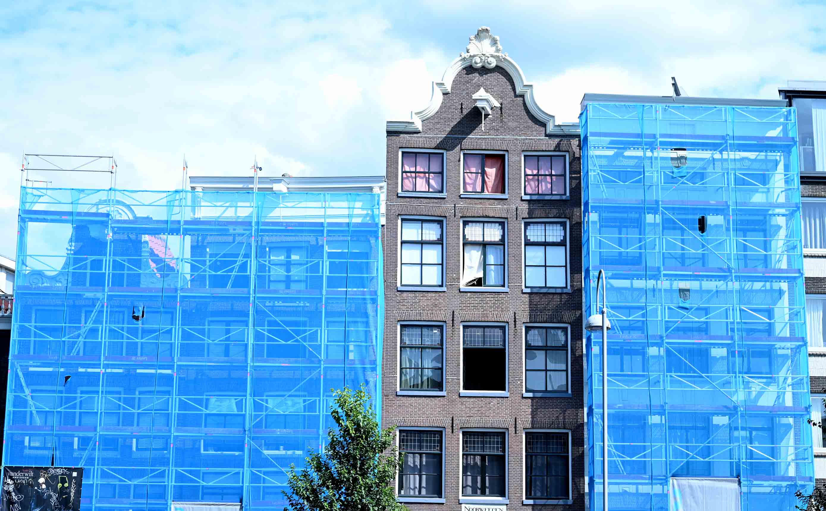Steigers met blauw stofnet rond grachtenpandgevels in Amsterdam tijdens verbouwwerk.