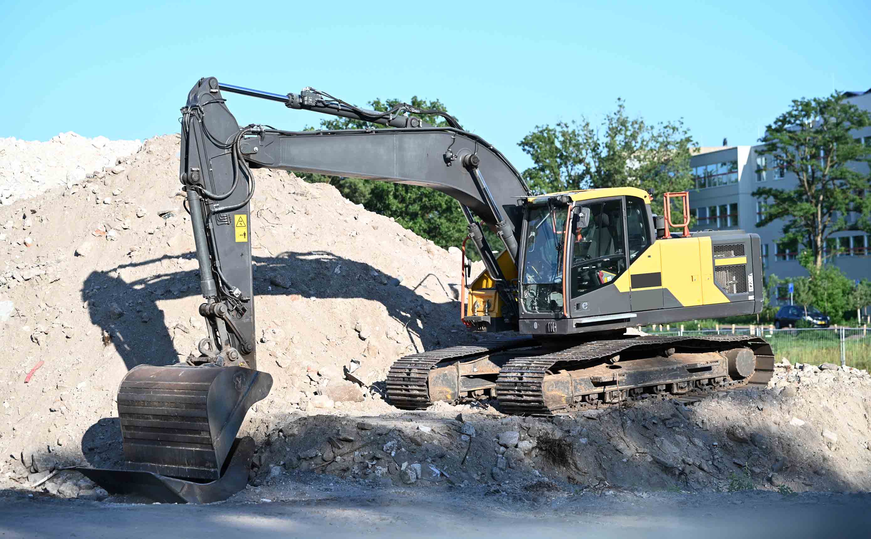 Een rupsgraafmachine werkt aan een berg puin op een Nederlandse bouwplaats, met een woongebouw op de achtergrond.