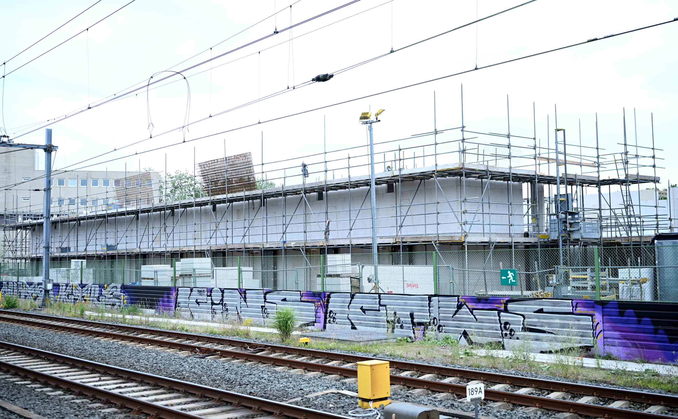 A concrete structural frame under construction beside a railway line, wrapped in scaffolding, with precast floor planks and a mat of reinforcement steel waiting on the top deck.