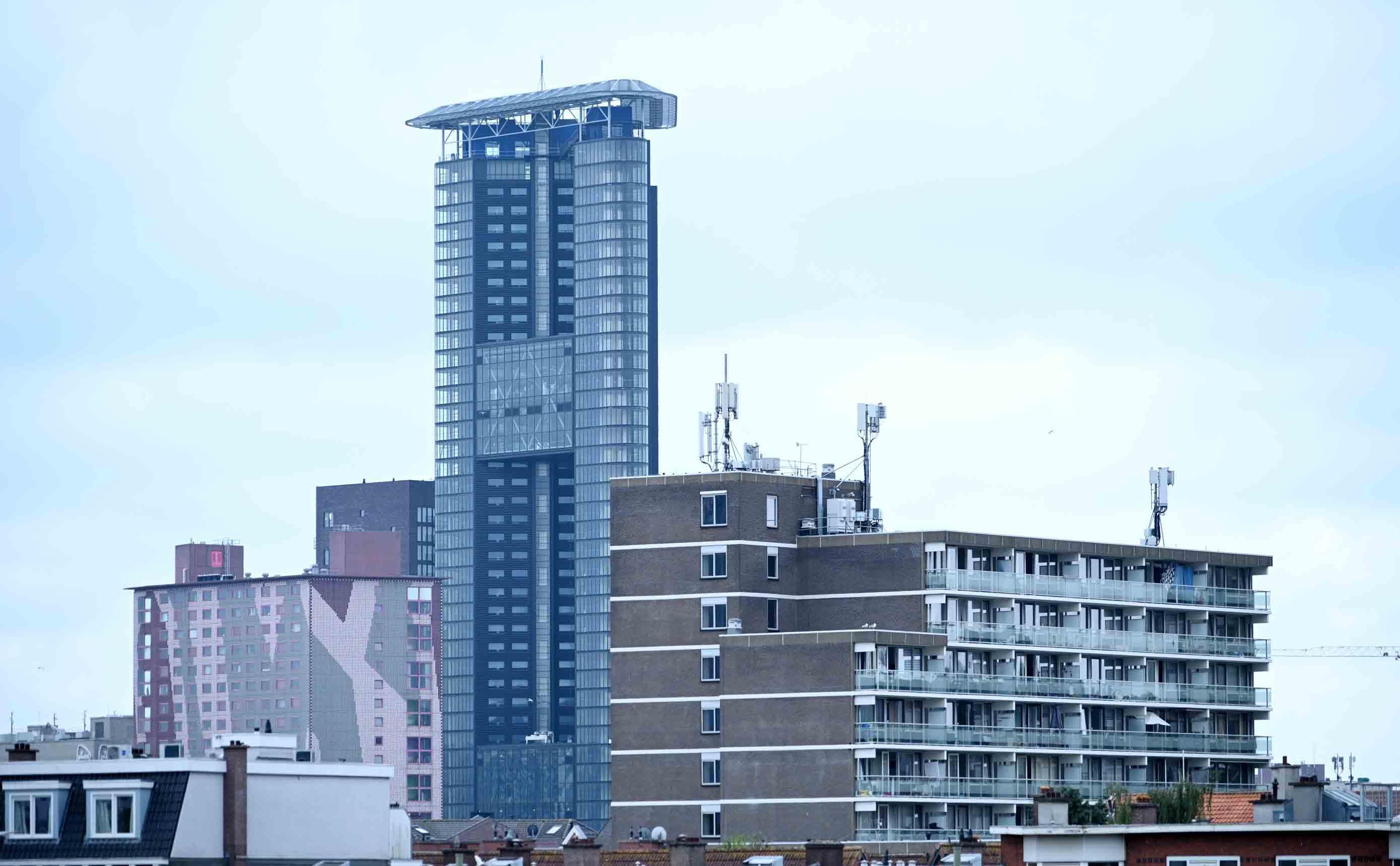 A Dutch city skyline of finished residential and office towers, older brick blocks standing beside a tall glass tower, all in use under a pale sky.