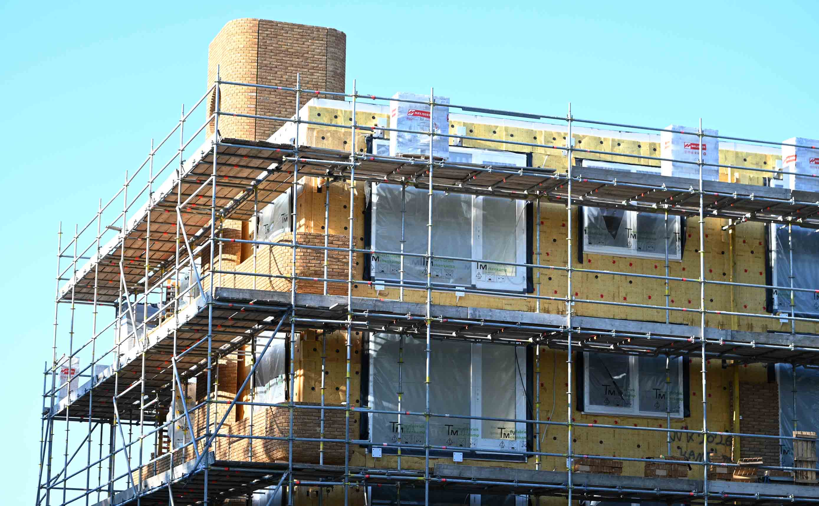 Een woongebouw in aanbouw, in de steigers, met gele minerale-wolisolatieplaten en een dampscherm zichtbaar op de gevel achter een bakstenen buitenblad.