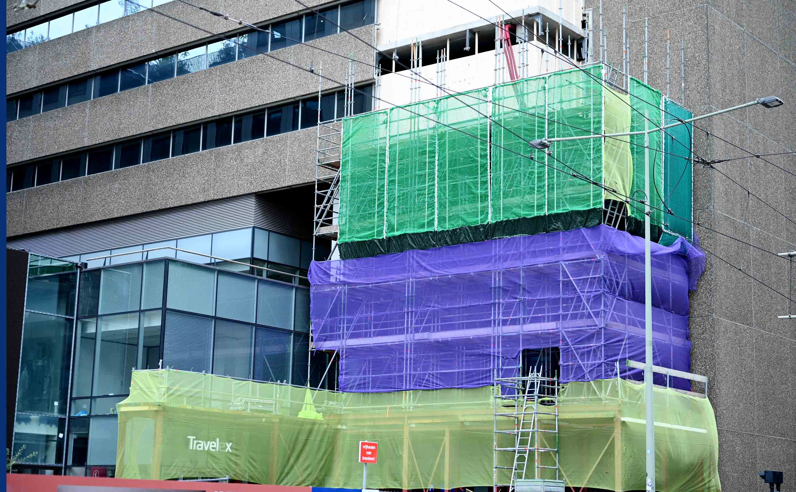 Steiger gehuld in groen, paars en geel veiligheidsnet tegen een gevel in renovatie, met toegangsladders tussen de lagen.