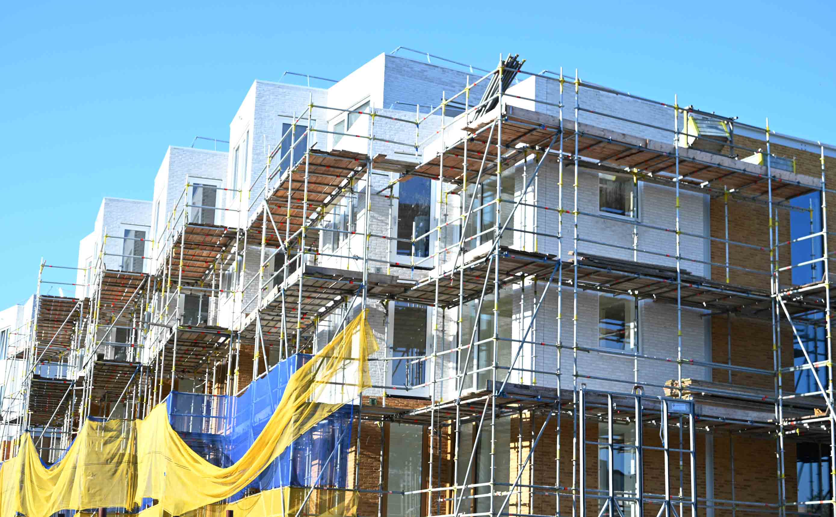 Een woongebouw in aanbouw in de steigers, gehuld in blauwe en gele veiligheidsnetten, met een deels afgewerkte bakstenen gevel en ramen onder een heldere lucht.