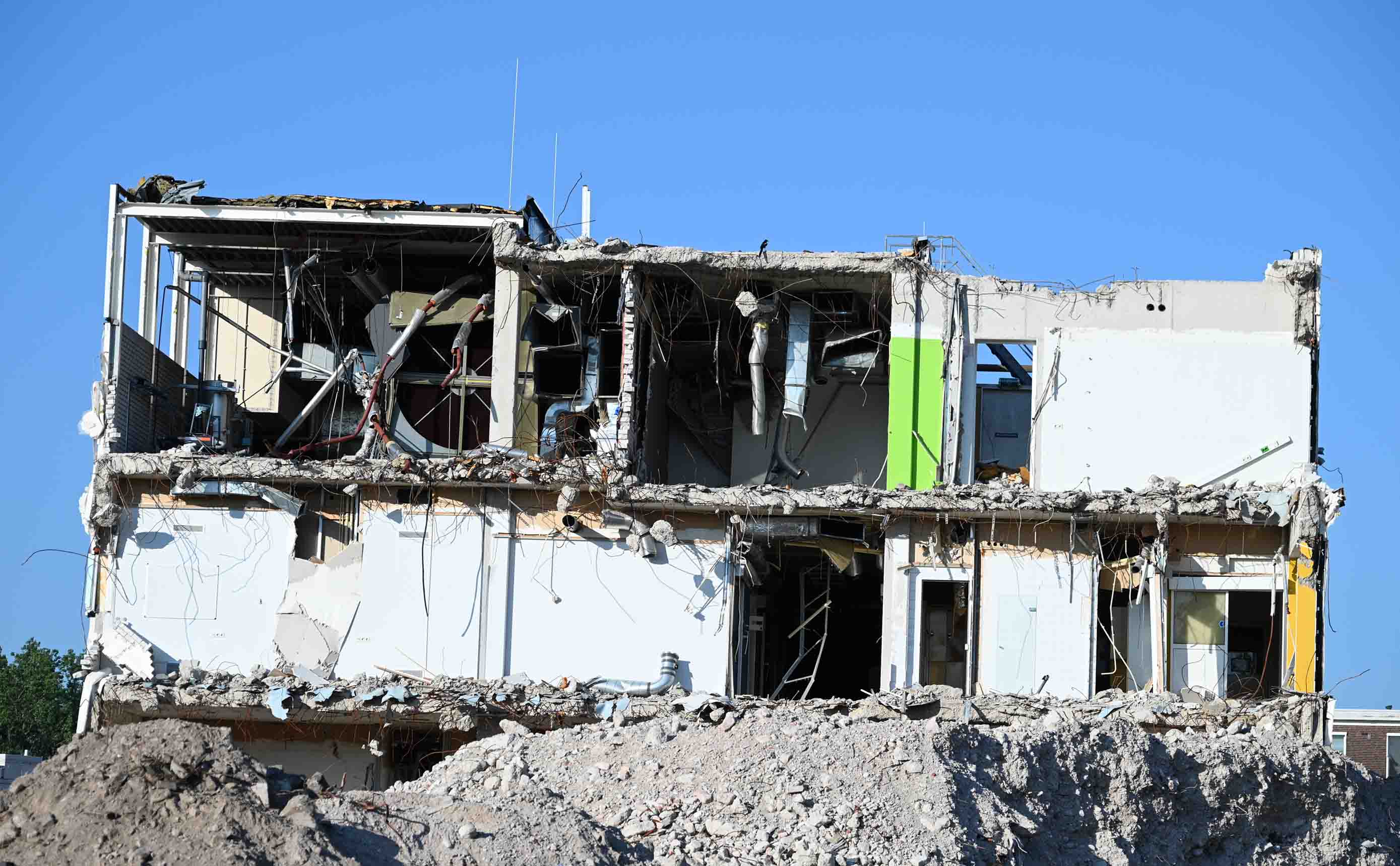 Een gebouw in doorsnede tijdens de sloop, met twee verdiepingen betonvloeren, binnenwanden en blootliggende ventilatiekanalen en installaties tegen een blauwe lucht.