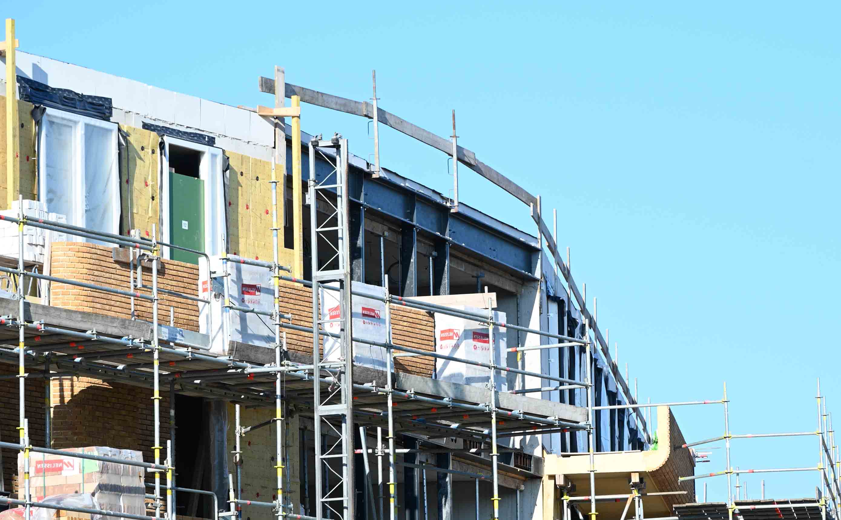 A facade under construction wrapped in scaffolding, with steel columns, a brick outer leaf, mineral-wool insulation and open window reveals all visible at once.